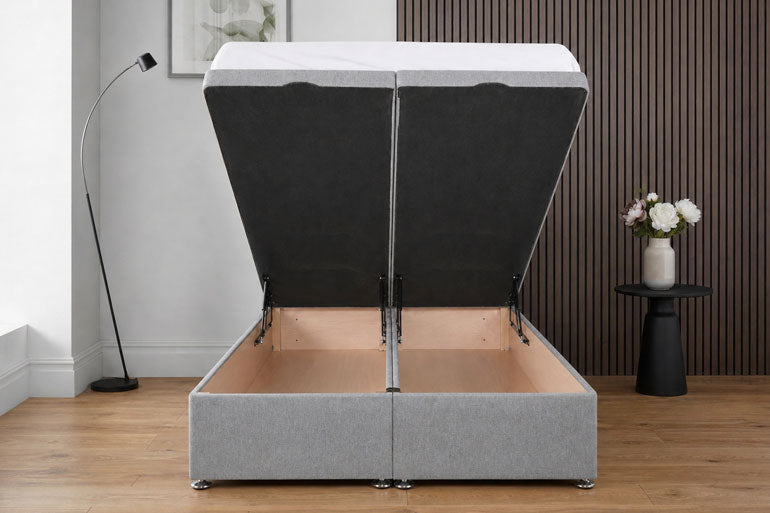 A double end lift ottoman bed in light grey, shown open to reveal a light wood interior. The bed is set in a modern bedroom with a wood slat wall, a floor lamp, and a small side table with flowers.
