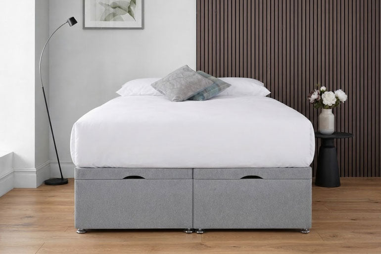 An end lift ottoman storage bed with a white mattress and pillows, shown in a modern bedroom setting with a dark wood slatted wall, floor lamp, and side table.