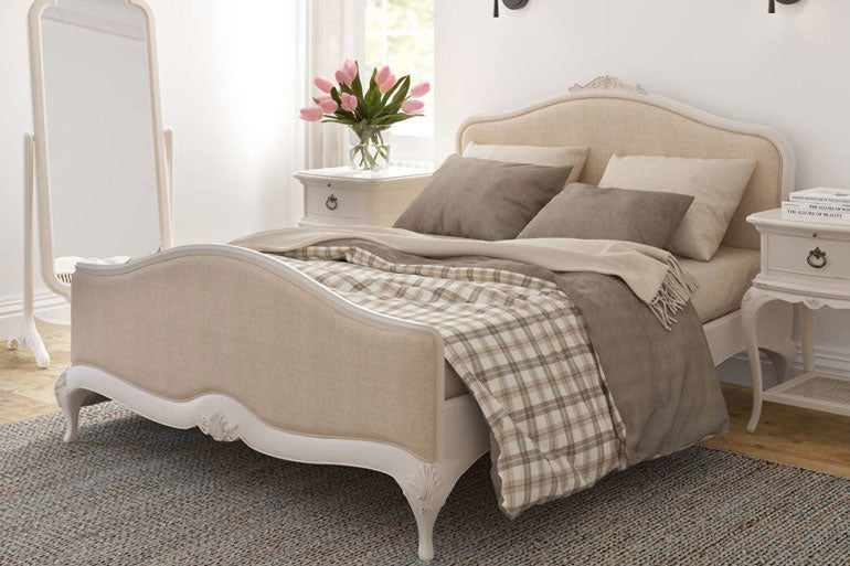An ivory upholstered bed with a high headboard and footboard, featuring hand carvings and a soft painted finish, placed in a bedroom setting with bedding and a bedside table.