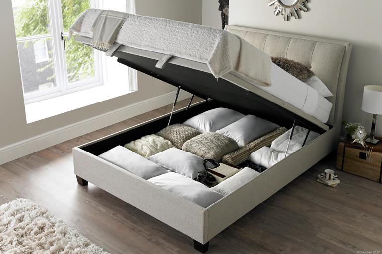 Kaydian Accent Ottoman Bed in Oatmeal with storage compartment open and bedding inside.