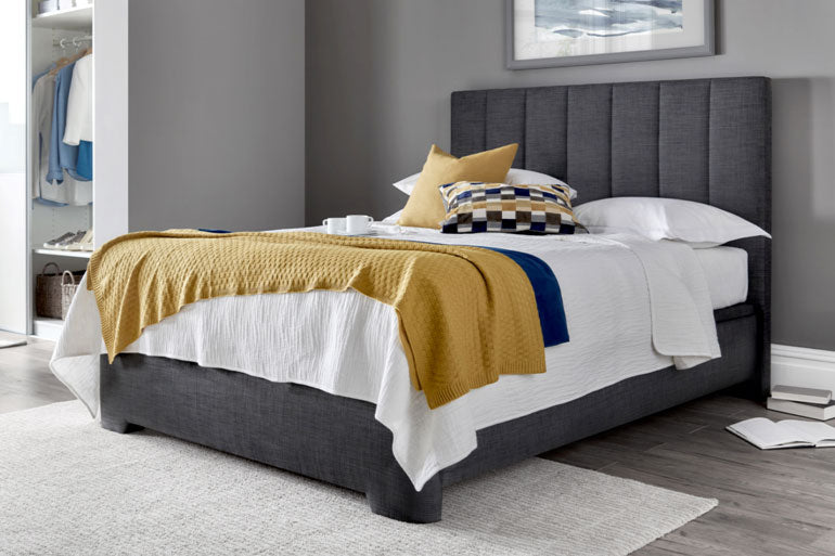 A Kaydian Medburn Ottoman Bed in Slate with white bedding and yellow throw and cushions.