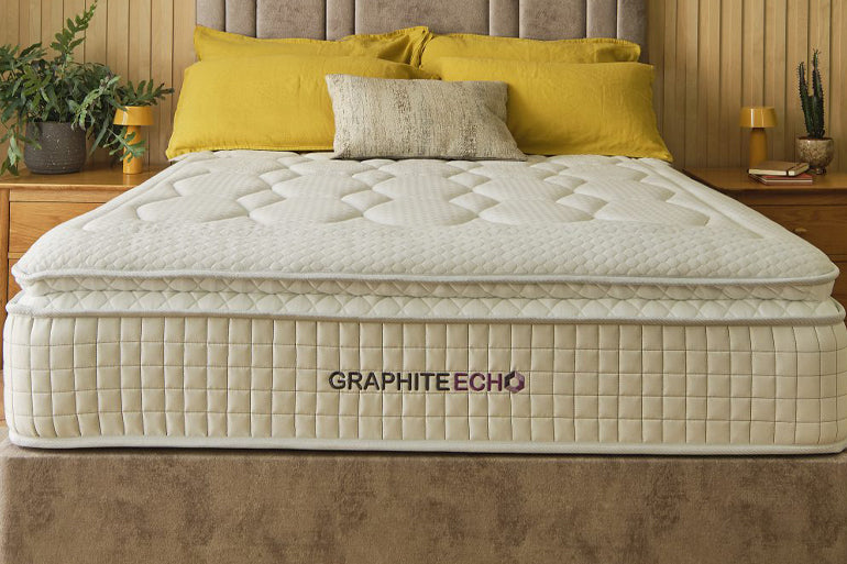 White mattress with 'Graphite Echo' branding on a bed with yellow pillows and fabric headboard.