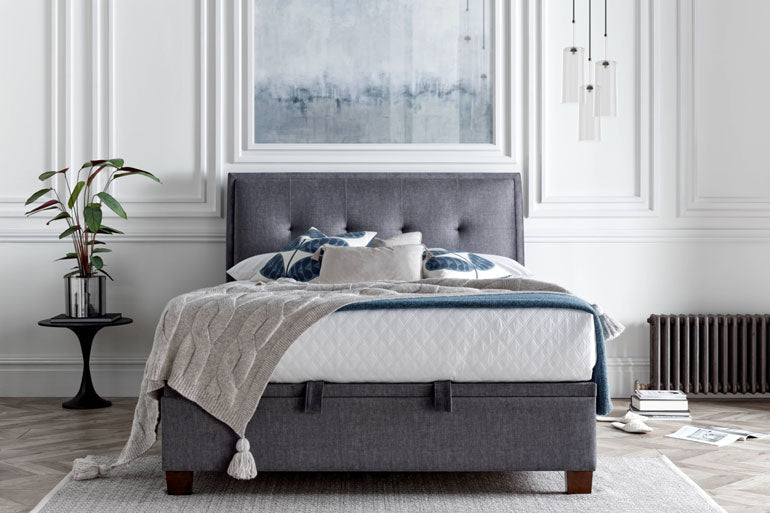 Kaydian Accent Ottoman Bed in Vogue Grey with a neatly made bed with a white beddng, in a room with a light decor.