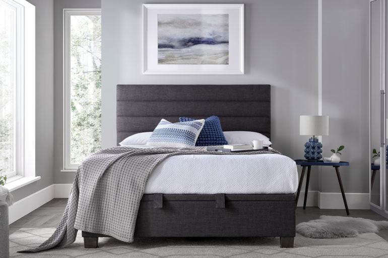 Kaydian Appleford Ottoman Bed in a bedroom with a side table and lamp.