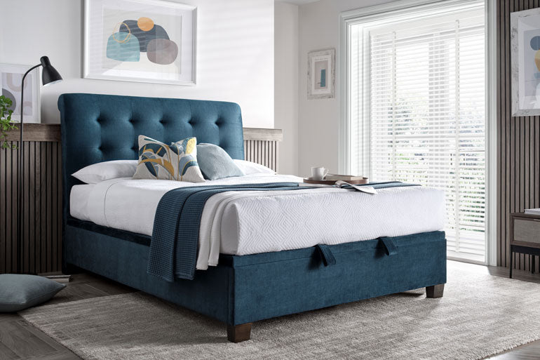 Kaydian Ashbrooke Ottoman Bed in Vogue Ocean Blue in a bedroom with white bedding and decorative pillows.