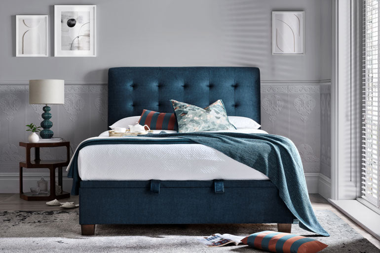 Kaydian Ashbrooke Ottoman bed in Vogu Blue a modern bedroom with gray walls and a lamp.