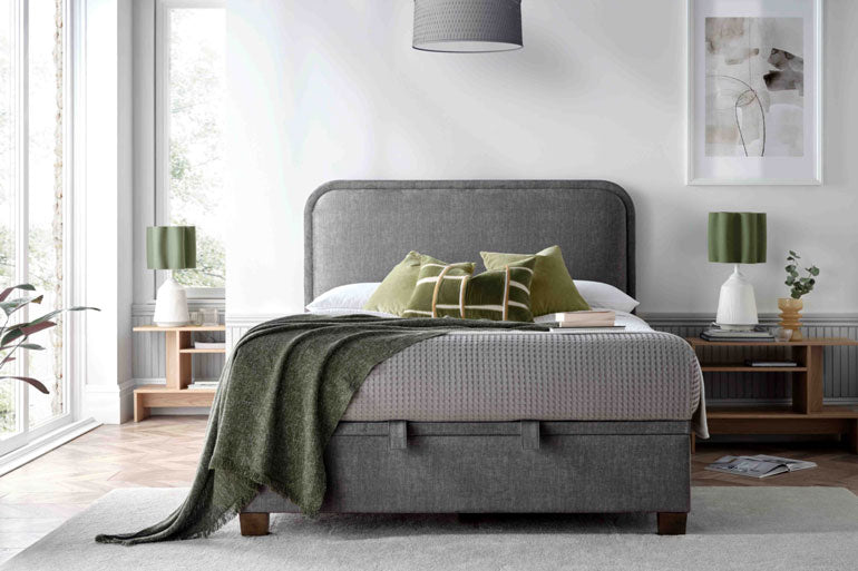 A grey upholstered Kaydian Clifton Ottoman Bed with a high headboard, low foot end, and wooden legs in a bedroom setting, featuring decorative pillows and a throw blanket.