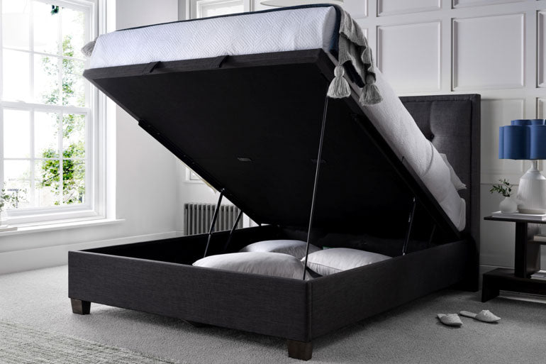 Kaydian Falfield Ottoman Bed in slate with storage compartment in a room with a window and lamp.