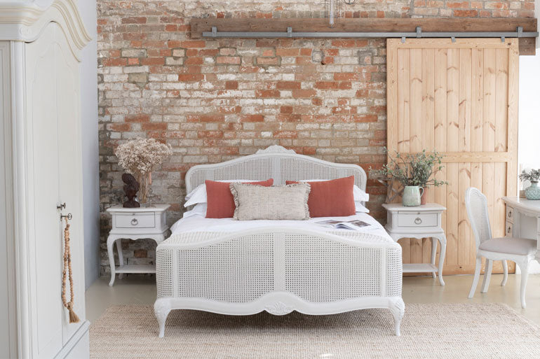 An elegant, gray-colored rattan high-end bed with a French style headboard and footboard, placed in a room with a wooden armoire and a white rug.