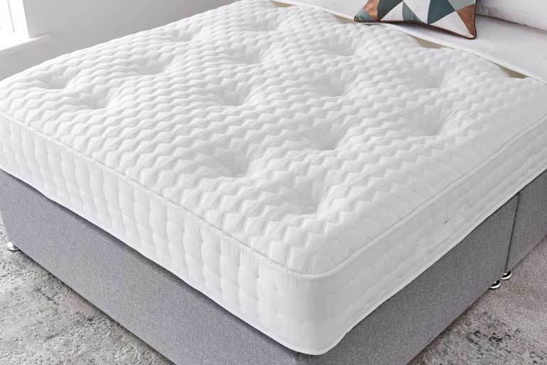 A white Giltedge Bliss 1000 mattress, placed on a bed base with decorative pillows at the head.